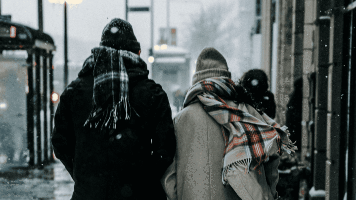 To personer går på gaden iført hue og store halstørklæder - der er is og sne på fortovet.