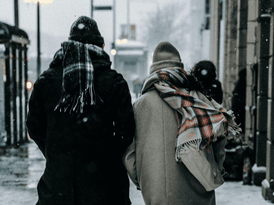 To personer går på gaden iført hue og store halstørklæder - der er is og sne på fortovet.