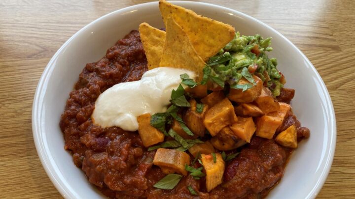 Chili sin carne serveret med tortillachips, cremefarice og hakket grønt i en hvid dyb tallerken.
